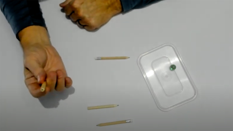 Hands on a table holding a short pencil, there is also a plastic tray and marble on top of it.